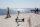 A photograph of a scooter parked on a cobblestone surface besides the sea, there are people standing on either side and a child cycling a bike. In the distance is a plane and mountains.