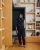 A portrait of Nikila Srinivasan at Meta's office in New York wearing a black sleeveless top and tailored pants, with red heels. She poses in a niche surrounded by wooden bookshelves filled with books and decorative items.