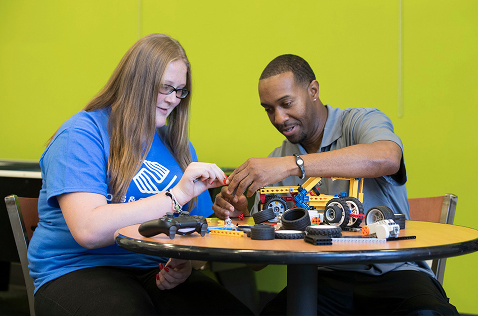 A young woman and young man working on a robotics project inside a classroom.