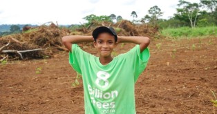 Young kid standing in the middle of an open field wearing an 8 Billion Trees brand green t-shirt raises his hands after a tree planting education session aimed at teaching kids the importance of calculating emissions in the central Amazonian State of Brazil