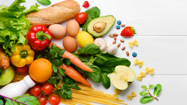 An assortment of fresh, colorful fruits and vegetables including lettuce, tomatoes, avocado, carrots, peppers, eggs, nuts, and berries, arranged on a white wooden surface.