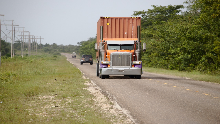 A large orange container truck driving down a rural two-lane road with greenery on the sides and other vehicles in the distance.