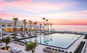 a large outdoor pool is surrounded by lounge chairs and palm trees , with the ocean in the background at Grecotel LUXME White