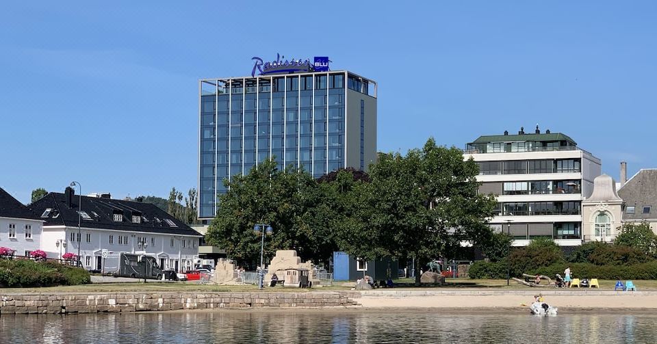 "a tall building with the logo "" radisson blu "" on it , situated near a body of water and surrounded by trees" at Radisson Blu Caledonien Hotel, Kristiansand