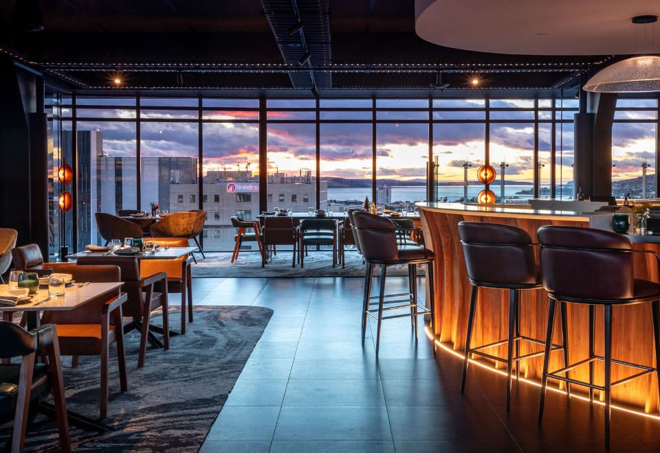 a modern bar with a large window , wooden table , and chairs , and the sun setting in the background at Crowne Plaza Hobart, an IHG Hotel