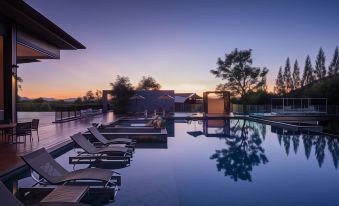 a large outdoor swimming pool surrounded by lounge chairs , with the sun setting in the background at dusitD2 Khao Yai