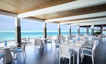 a large , modern restaurant with white tables and chairs arranged on a patio overlooking the ocean at Anantara Dhigu Maldives Resort