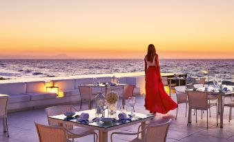 a woman in a red dress standing on a rooftop patio with a beautiful ocean view , enjoying the sunset at Grecotel LUXME White