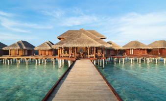 a wooden pier leads to a thatched - roof bungalow with clear blue water in the background at Anantara Dhigu Maldives Resort