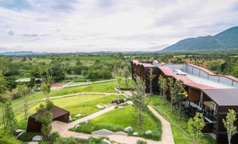 a large wooden building with a red roof is surrounded by green grass and trees , with a mountainous landscape in the background at dusitD2 Khao Yai