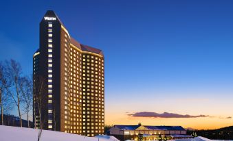 a tall hotel building surrounded by snow - covered mountains , with the sun setting in the background at The Westin Rusutsu Resort