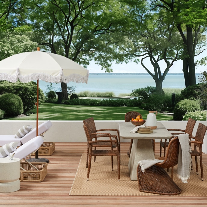 White lounge chairs under a white umbrella and a woven wicker outdoor dining set on a patio with a lake-side view.