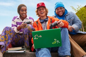 a trio of colorfully dressed students sitting side by side holding video game controls and playing a game on a laptop. 