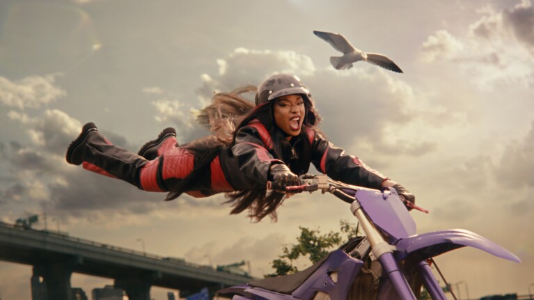 An image of Megan Thee Stallion holding onto the handlebars of a motorbike while she soars through the air in a stunt.