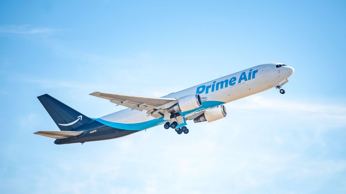 An image of a plane in flight. The decals on the plan say "Prime Air" in blue lettering with a light blue and navy blue design around the tail and the belly. There is a white Amazon smile logo on the tail of the plane.
