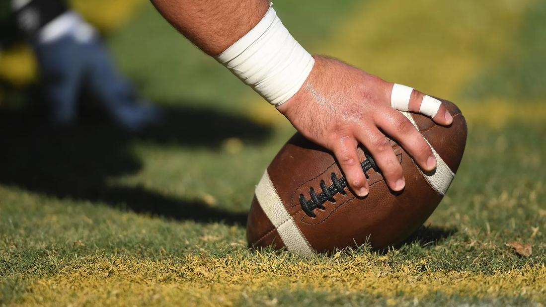 Large hand with athlete-taped wrist and fingers gripping football on the field