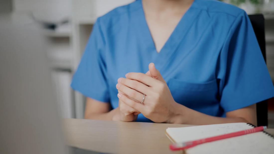 Person in scrubs with hands folded, across from person at desk