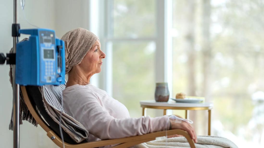 Person receiving cancer infusion therapy sitting in chair, head wrapped in scarf, gazing out window