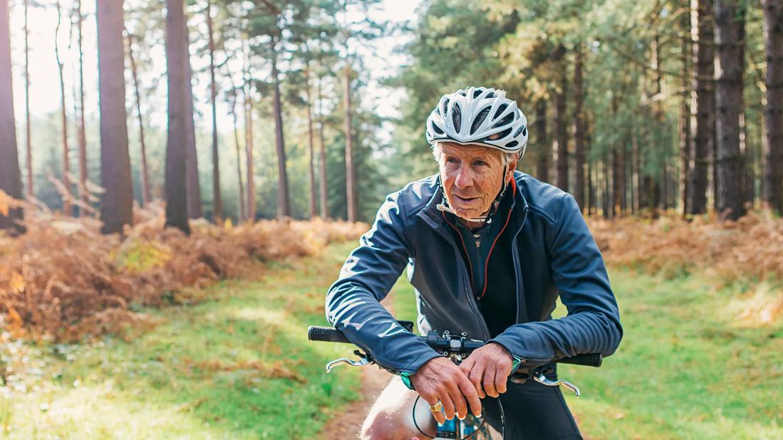 Older person, in bike helmet, sitting on bike on trail in woods taking a break