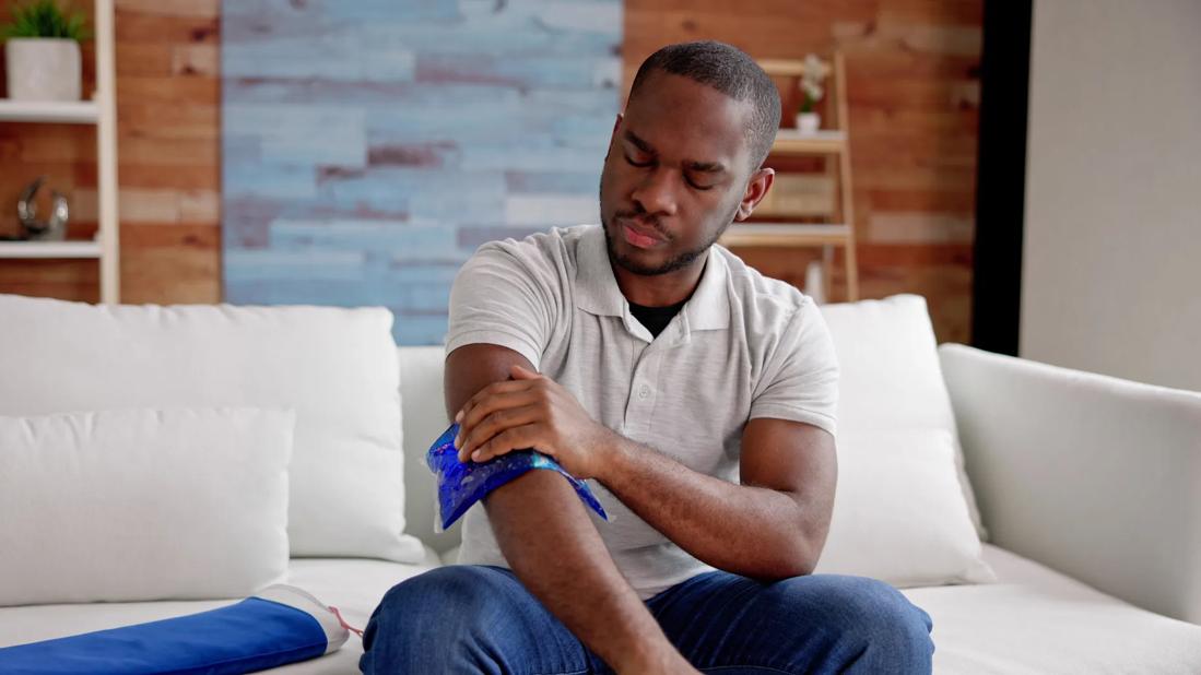 Person sitting on couch applying cold compress to arm, with heating pad nearby