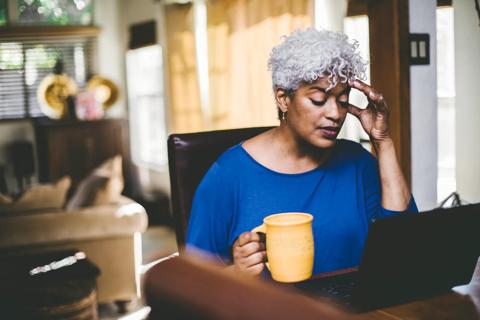 Female looking at laptop at home desk in living room, hand on head, holding coffee cup, looking stressed