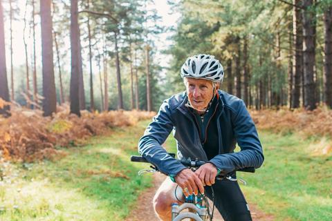 Older person, in bike helmet, sitting on bike on trail in woods taking a break