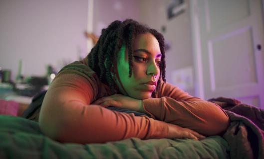 Person lying stomach down on bed, head on crossed arms, staring into the distance