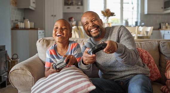 Happy, laughing child and caregiver sitting on couch holding video game controllers, playing video games