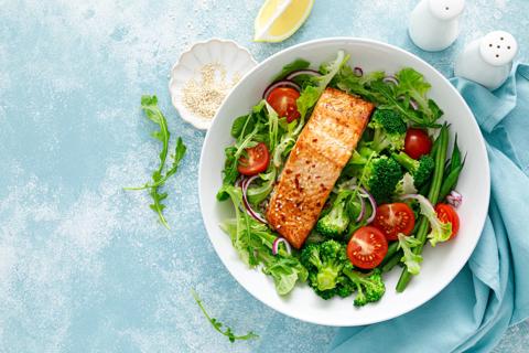 Plate filled with mixed greens and veggies and grilled salmon, with small bowl of sesame seeds and a slice of lemon on table