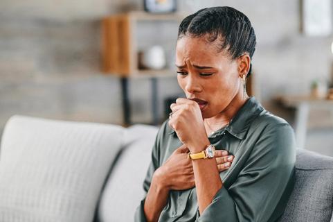 Person on couch, brows furrowed coughing into hand, with other hand on their chest