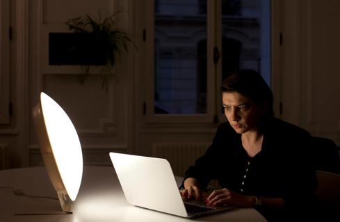 Person sitting at home desk working on laptop, with a light box for light therapy nearby