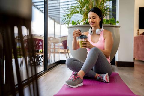 A person in exercise clothes sits on a yoga mat and holds a smoothie cup