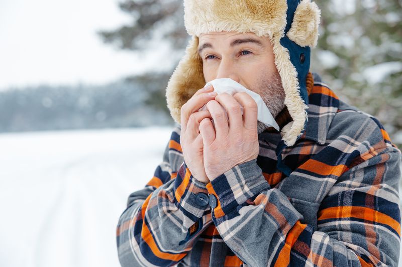 A man in a checkered coat and a fake-fur lined trapper hat is blowing his nose. He is standing in what looks like a snow covered field with a tree in the background. 