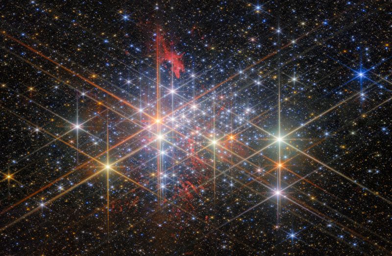 A dense cluster of bright stars, each with six large and two small diffraction spikes, due to the telescope’s optics. They have a variety of sizes depending on their brightness and distance from us in the cluster, and different colours reflecting different types of star. Patches of billowing red gas can be seen in and around the cluster, lit up by the stars. Small stars in the cluster blend into a background of distant stars and galaxies on black.]