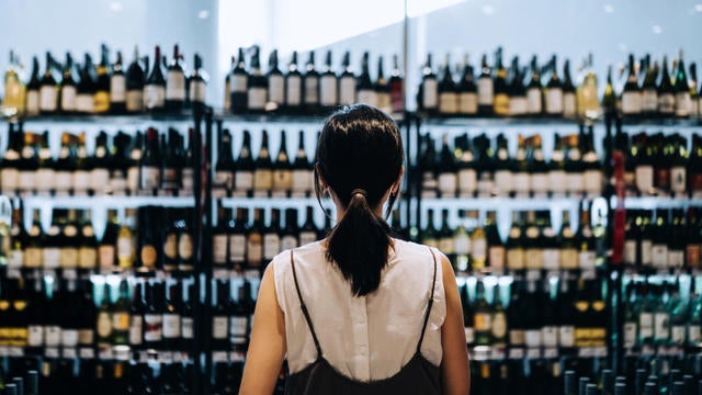 Rear view of young Asian woman grocery shopping for wines in a supermarket. She is standing in front of the liquor aisle and have no idea which wine to choose from 