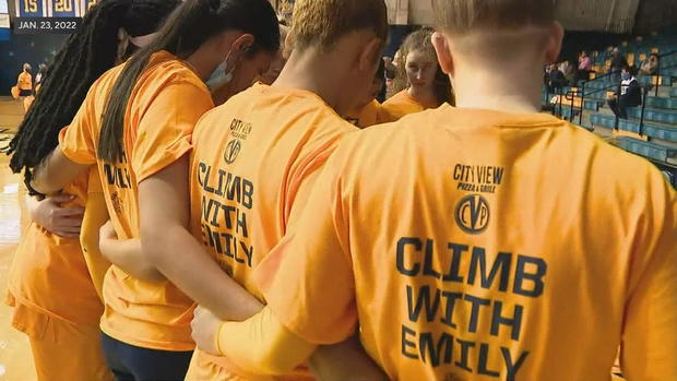 Women's basketball players in a huddle wear yellow t-shirts that say Climb with Emily on the back 