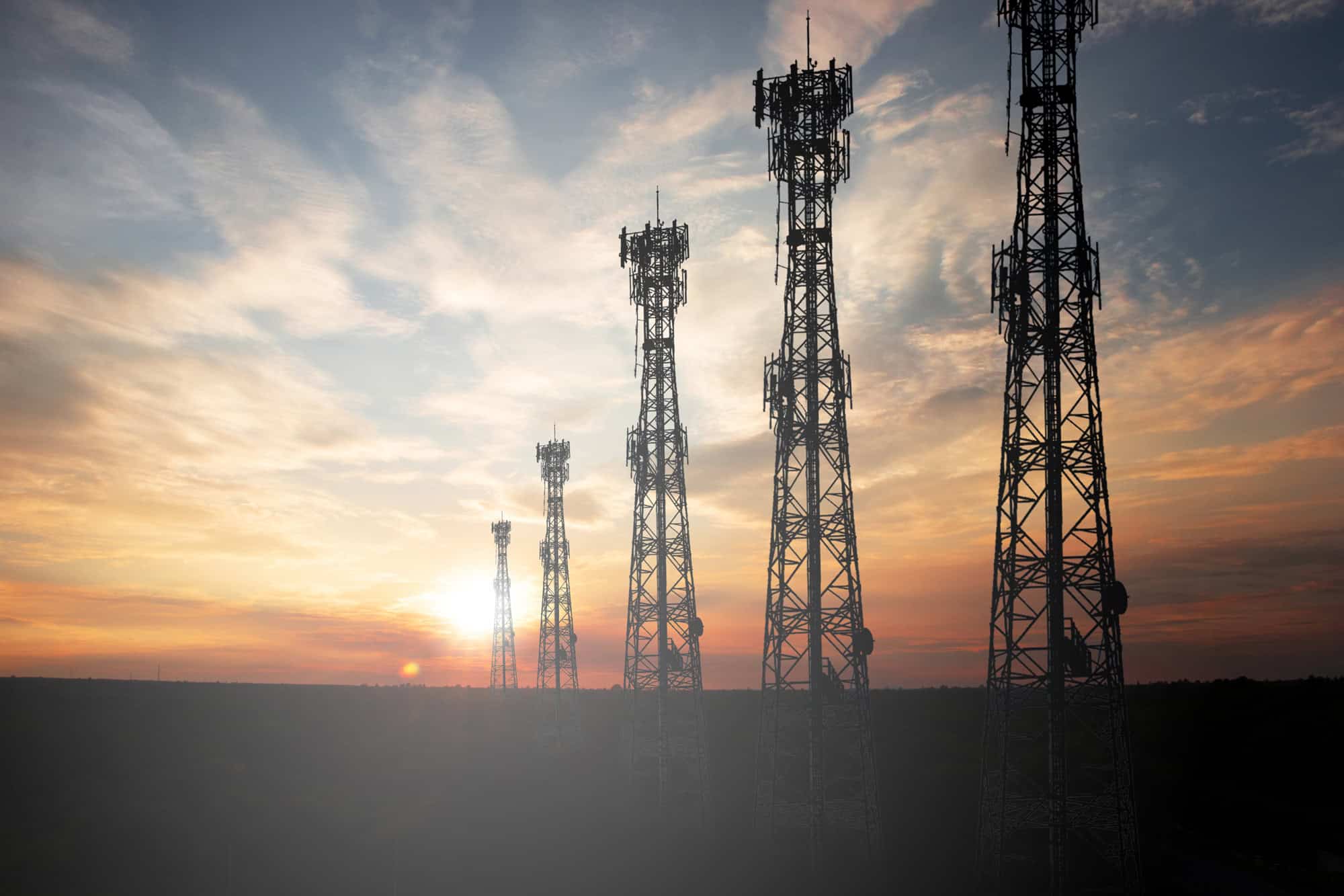 Five communication towers stand silhouetted against a sunset sky with scattered clouds.