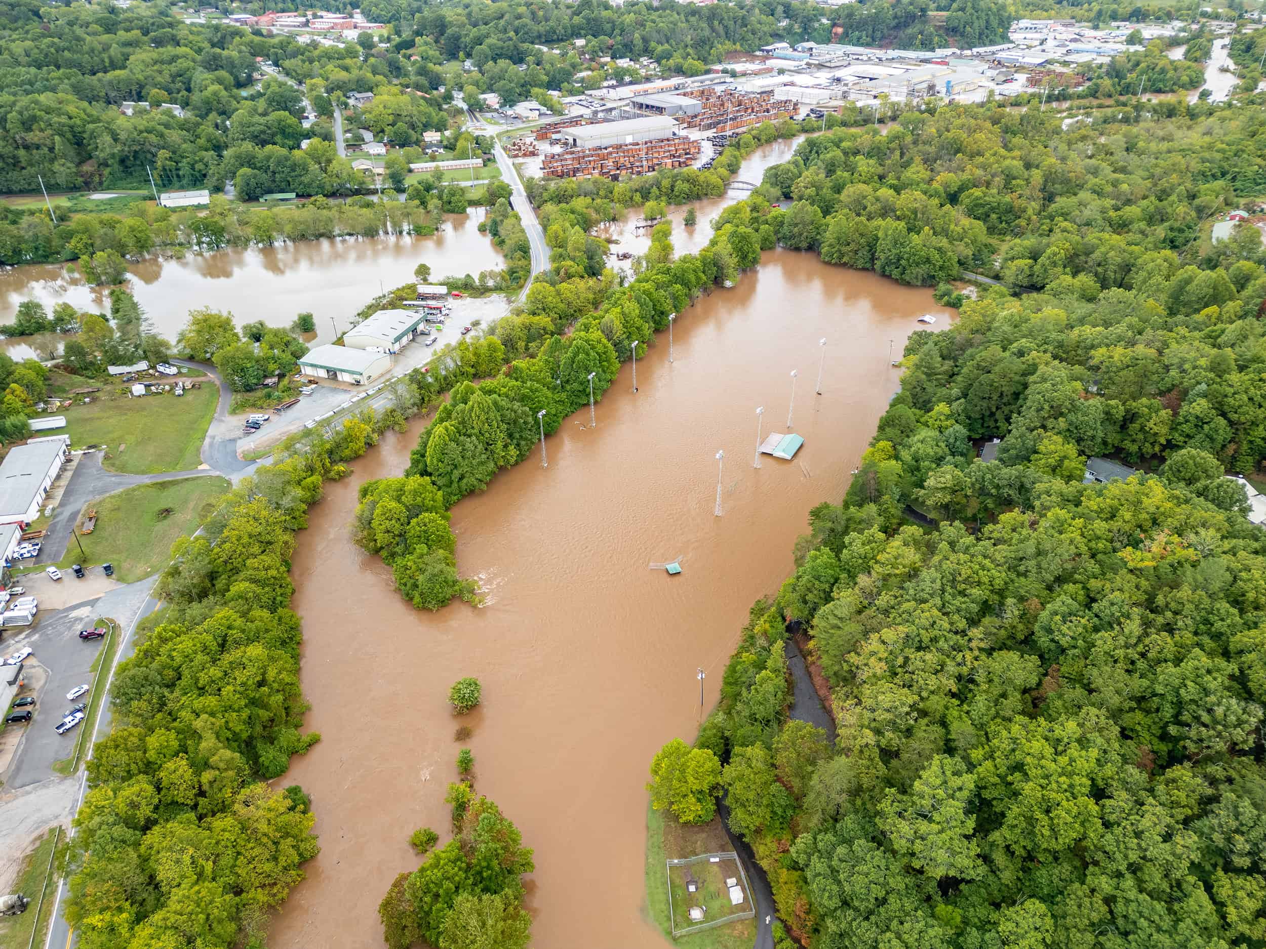 Aerial view of a flooded area in western North Carolina with muddy water, surrounded by trees and buildings. Soccer fields with visible goal posts are partially submerged.