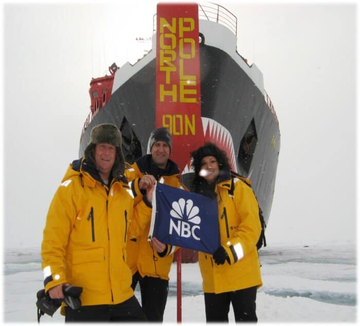 Three people in yellow jackets stand in front of a ship at the North Pole, holding an NBC flag.