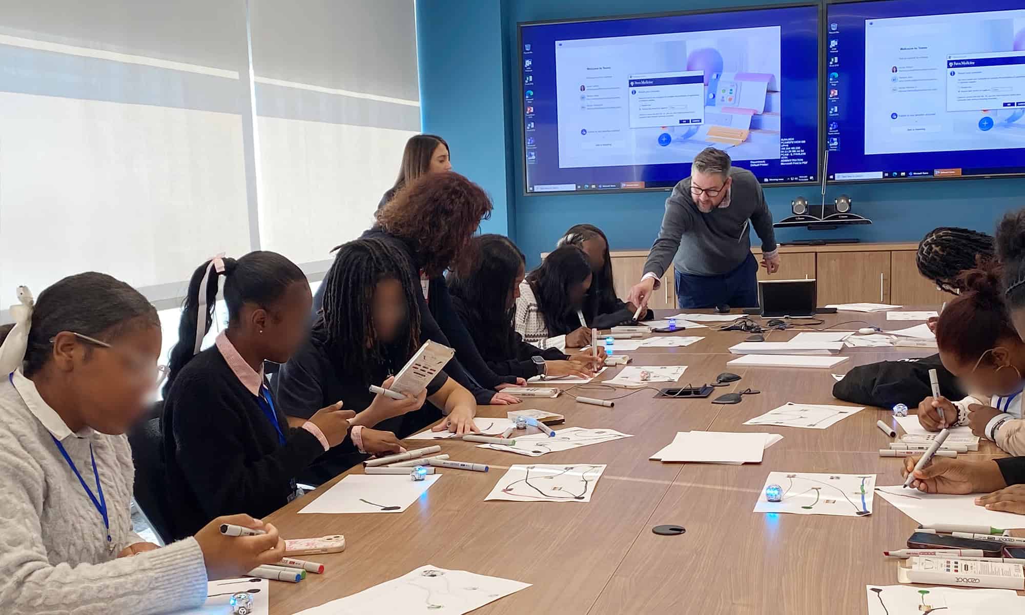 Group of people sitting at a conference table engaged in an activity with papers and electronic devices, while listening to a presenter pointing at a screen.