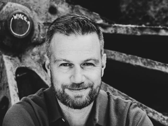 Black and white photo of a man with a beard smiling, set against a background with industrial machinery.