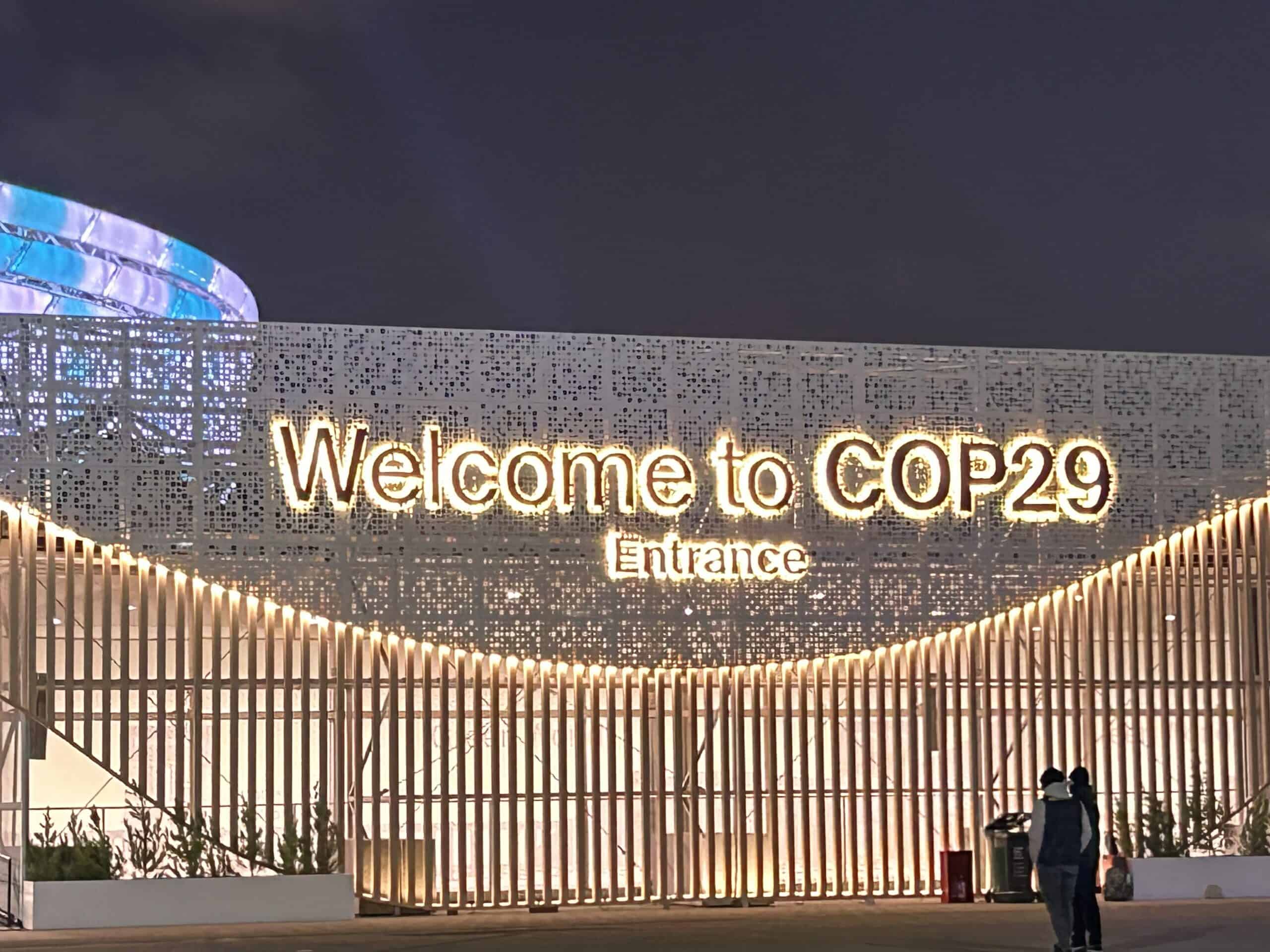 Sign with illuminated text reads "Welcome to COP29 Entrance" against a decorative backdrop at night.