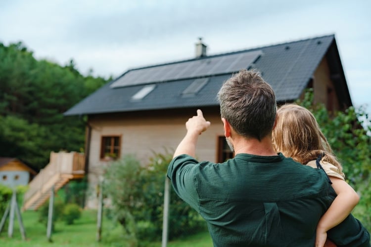 A man holding his daughter while counting the number of solar panels on their rooftop.