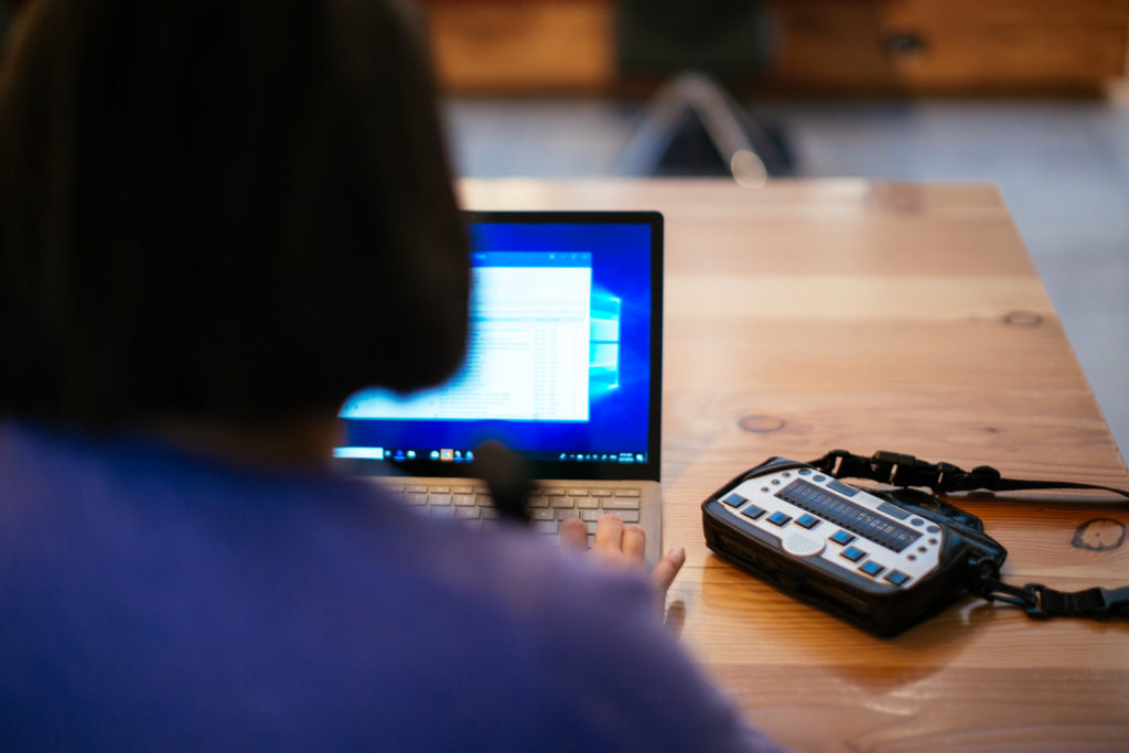 Anne Taylor seated at a table uses a laptop with a braille keyboard.