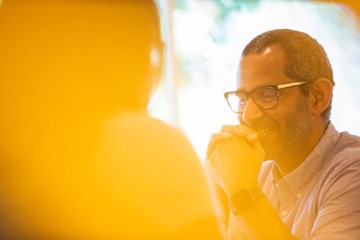 Picture of a middle aged man with glasses, hands near his mouth. There is a silhouette of a person in the foreground. 