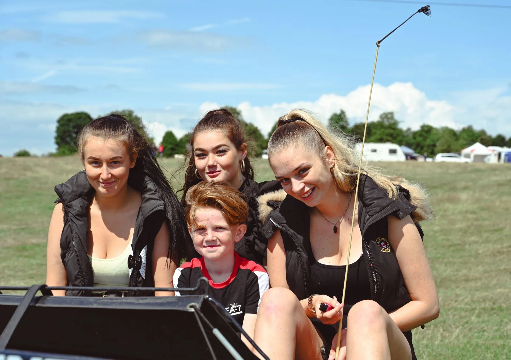 Image of 3 young women and a boy sat on the seat of a pony-trap. Green field in the background with blue skies