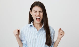 Studio portrait of angry and unsatisfied young european model showing fists and shouting with closed eyes, standing over gray ba