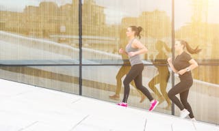 Two women running up a staircase with a big reflecting window on background. They are wearing gray and black sportswear. Healthy