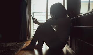 Asian woman sitting on wood floor beside window light in a dark room. Hold and bend head to look at mobile phone screen to check