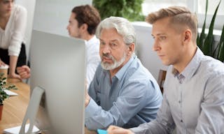 Focused senior and young colleagues discussing online project looking at pc screen, older mentor helps employee explaining compu